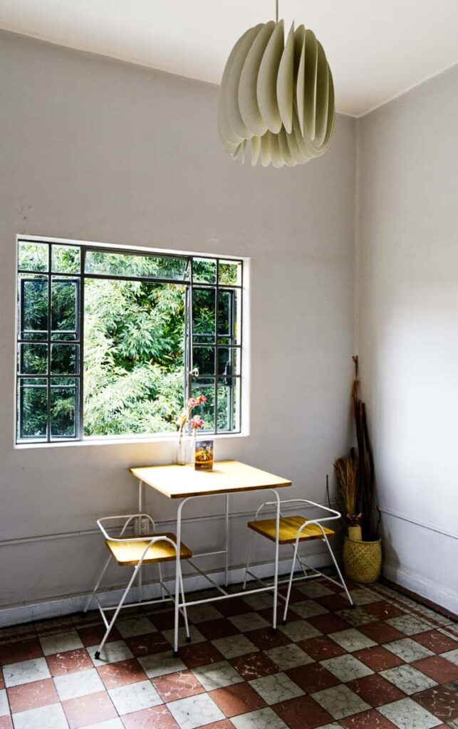 Modern decor characterizes Gluck Vegetarian Restaurant in Oaxaca. Two minimalist wooden table and chairs sits in front of a window with greenery as the view. A modern, deco paper lamp hangs from the ceiling.