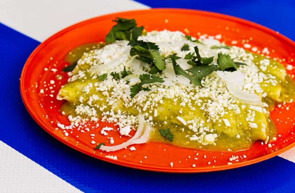 A plate of chicken enchiladas are rolled on a red plate and topped with a green salsa, fresh cheese, onions, and cilantro.
