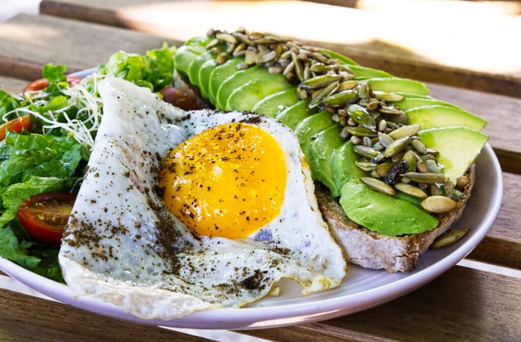 A plate of healthy breakfast in Puerto Escondido shows a piece of toast lined with large slices of avocado topped with sunflower and pumpkin seeds. On the side is a salad and sunny side up egg.