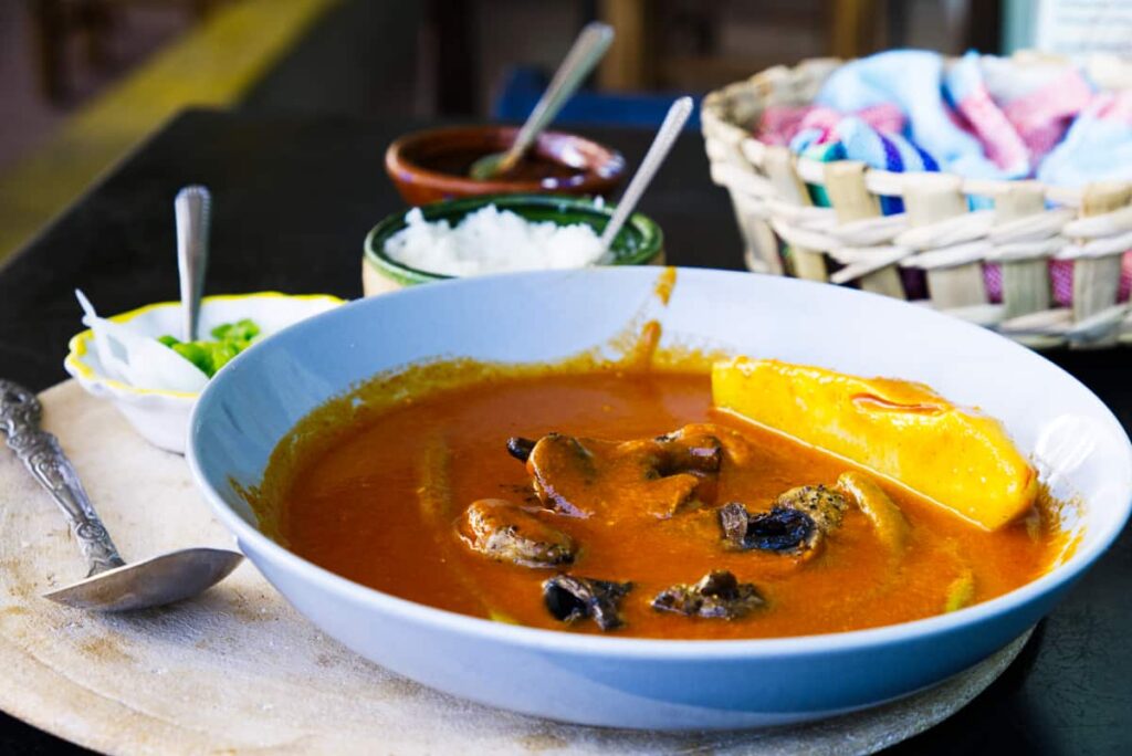 A round blue bowl filled with mushroom vegan mole sits on a table at Los Muchitos, one of the best vegan restaurants in Oaxaca. Surrounding the bowl are rice, chiles, salsa, and a basket of corn tortillas wrapped in a colorful fabric.