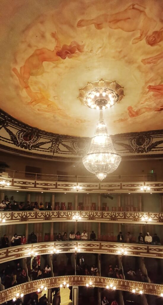 At the Macedonia Theater in Oaxaca, a large chandelier hangs from the ceiling which is painted with angelic like figures. Below is rows of box seating to watch a performance, one of the fun things to do in Oaxaca.