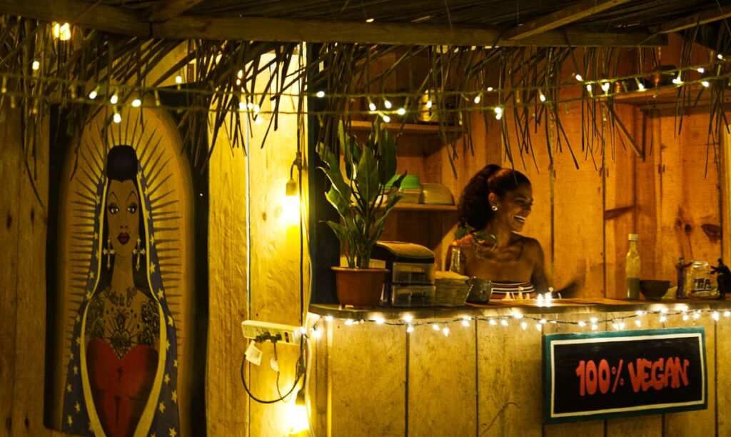 A woman smiles from behind the counter of her vegan restaurant in Puerto Escondido. The palapa stand features a sign that says 100% vegan and a large painted portrait of her on the right in the style of the Virgin de Guadalupe.