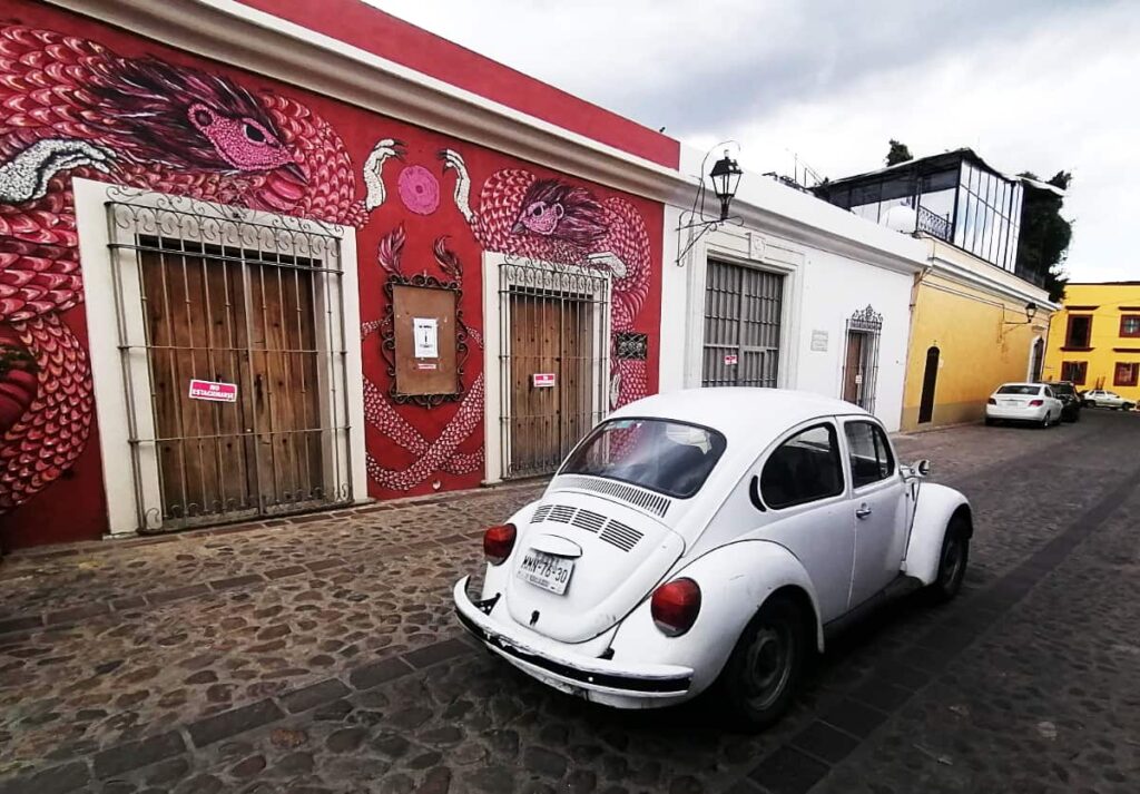 A white VW bug drives down a cobblestone street in Jalatlaco, one of the best places to find street art in Oaxaca. On the wall behind is a large red, pink, and white art of two bird-like figures.