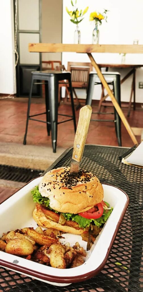 A vegan hamburger is served with a small knife sticking out of the bun. On the side are crispy potatoes. In the background are another table and chairs with vases of yellow flowers.