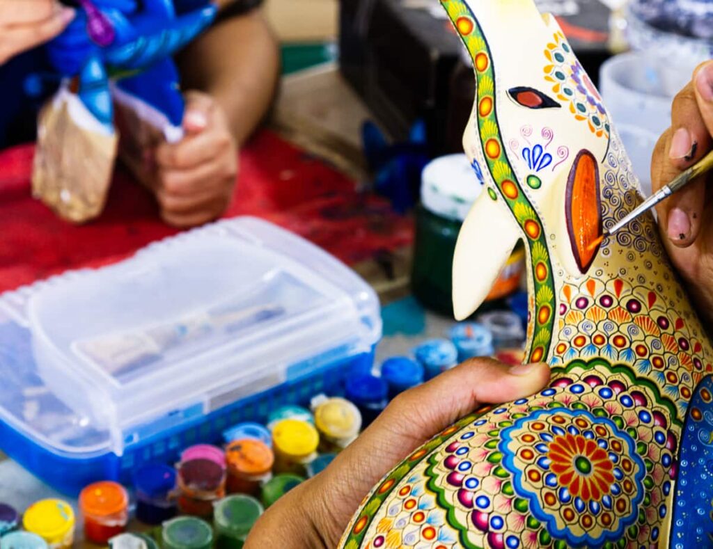 A hand paints an intricate alebrije, one of the unusual things to do in Oaxaca. In the background are several small containers of paint and another person painting an alebrije.