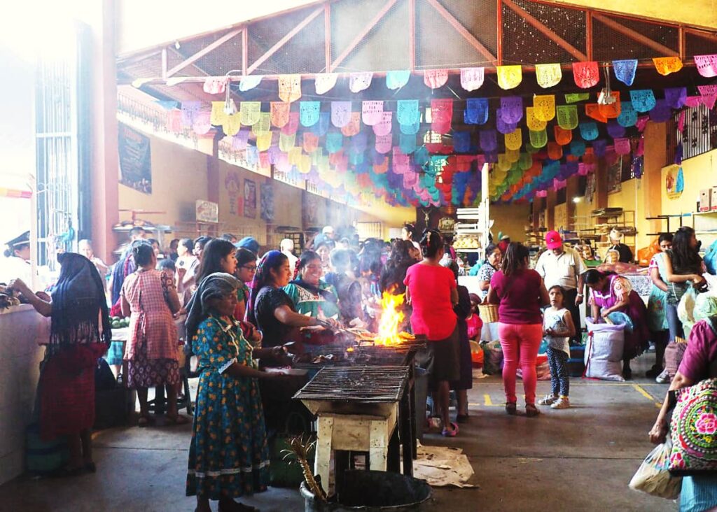 One of the top things to do in Oaxaca is explore the Tlacolula Market, home of the original smoke hall. In this image women dressed in traditional clothing cook meat and vegetables on the grills. In the background are more people and decorative flags hanging on the ceiling.