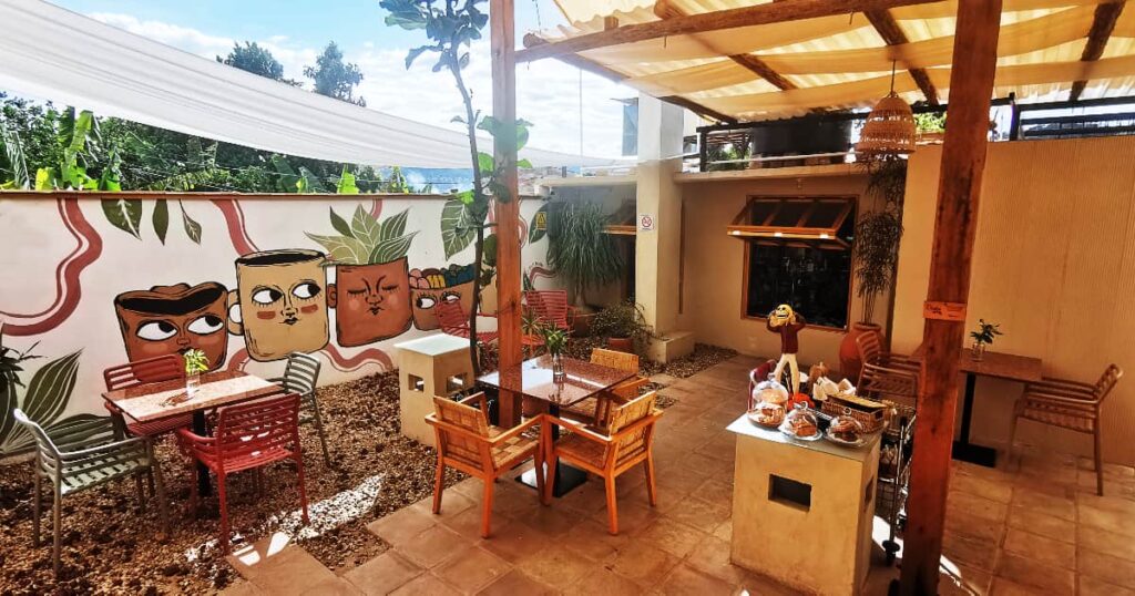 A shaded patio with wooden chairs and columns at a restaurant with vegan food in Oaxaca. The wall features a painting of several mugs with faces showing various expressions and surrounded by plants.