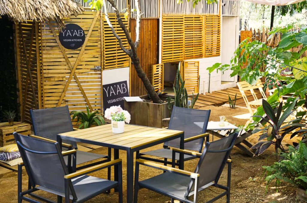 A garden view of one of the vegan restaurants in Puerto Escondido includes a table and chairs surrounded by tropical plants. In the background are two signs that read YA'AB Vegan Kitchen.