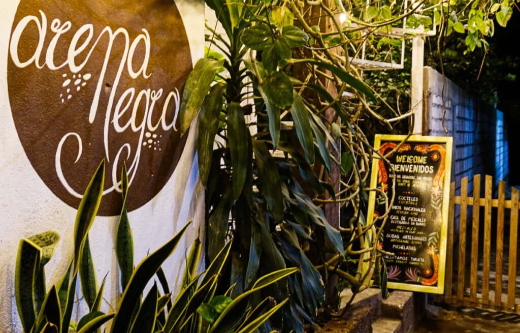 Outside of a Mazunte restaurant, a menu sign says Welcome in English and Spanish. On the wall is a painted logo with the words Arena Negra in white lettering on a brown background.