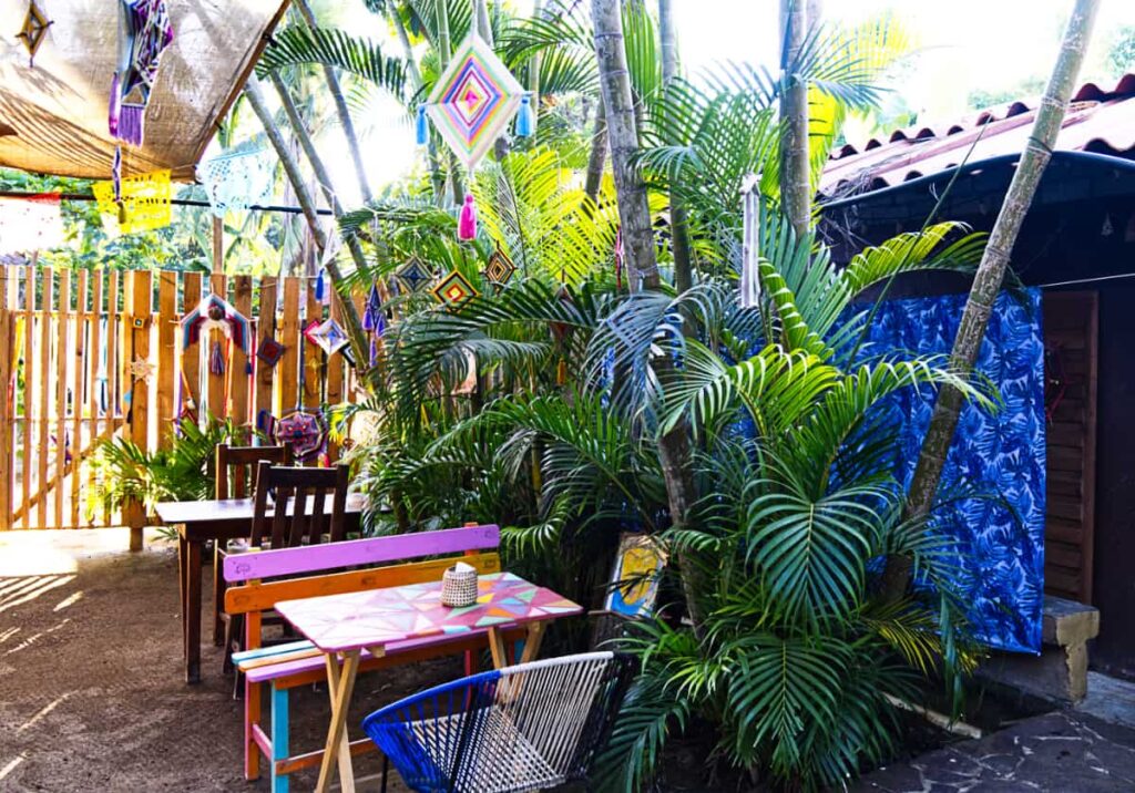 Tables sit outside, next to tropical plants at a cafe and bakery in Mazunte. Above, colorful decorations hang from the plants.