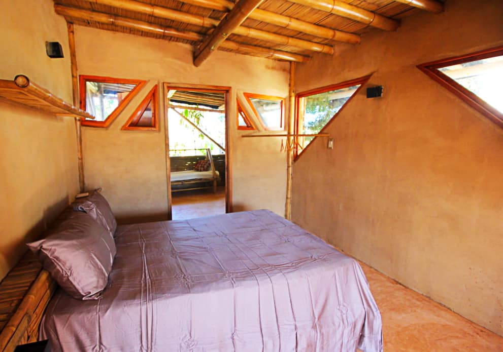 Inside a private bedroom at Bambu Corner Coliving in Puerto Escondido is a bad made with purple sheets. The walls are finished in mud with triangular shaped wooden framed windows. The ceiling and furniture are made of bamboo.