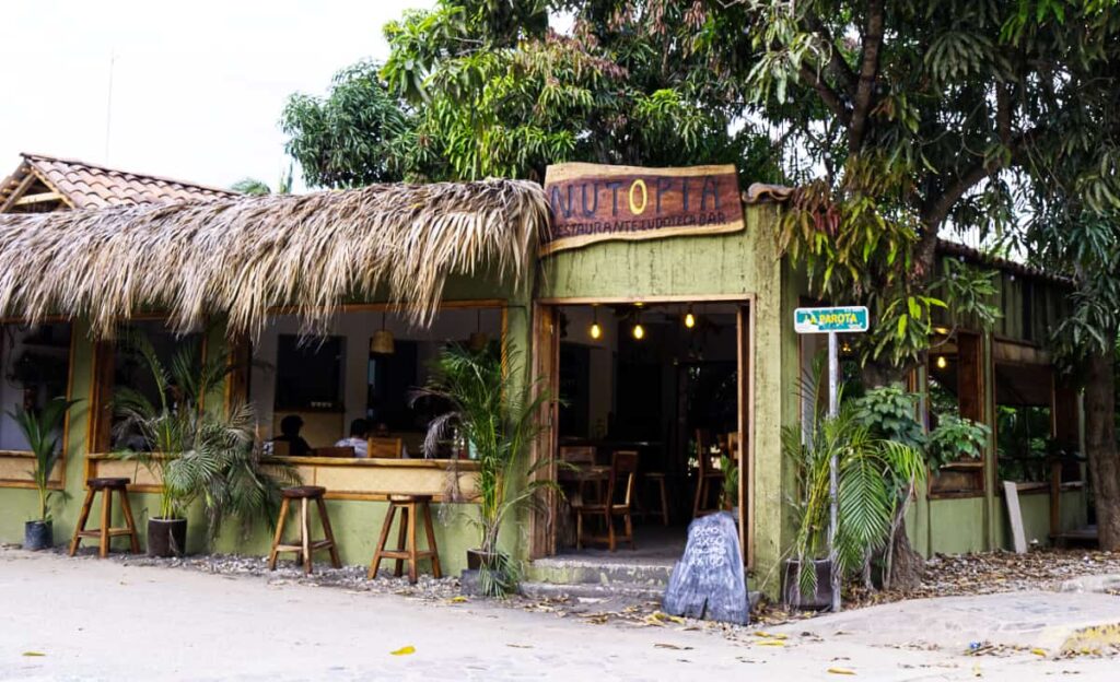 An outside view of a Mazunte restaurant. The green wooden building features wide open windows with a palapa roof. A few stools are outside and a wooden sign above the door says Nutopia, one of the best restaurants in Mazunte Oaxaca.