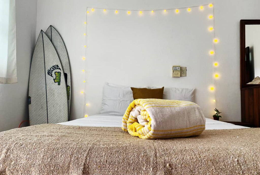 Inside a private bedroom at Casa Flow Coliving in Puerto Escondido, a bed is made up with a rolled towel sitting at the end. In the corner are two surfboards propped up against the wall. And yellow string lights hang on the wall above the bed.
