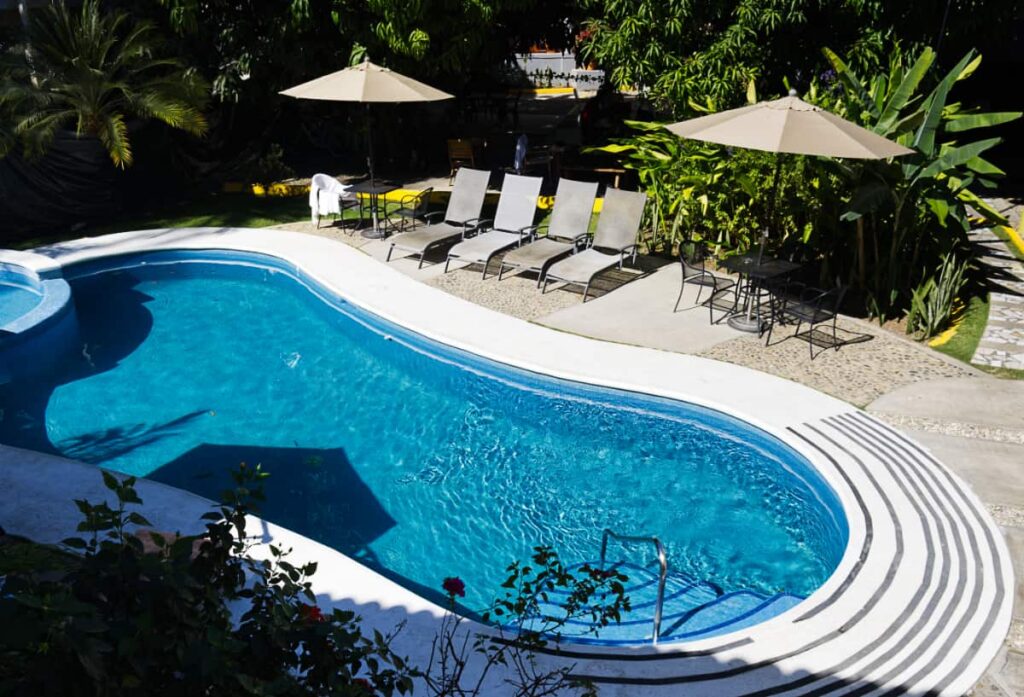 The blue, kidney shaped pool at Casa Losodeli is surrounded by sun loungers, two tables with an umbrella, and lots of plants. On the right, a paved path leads from the hostel area to the coworking space.