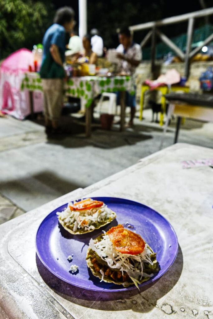 A night shot shows a purple plastic plate of two tostadas at the best place for cheap street food in Mazunte. In the blurred background, a man orders from the owners standing behind a table.