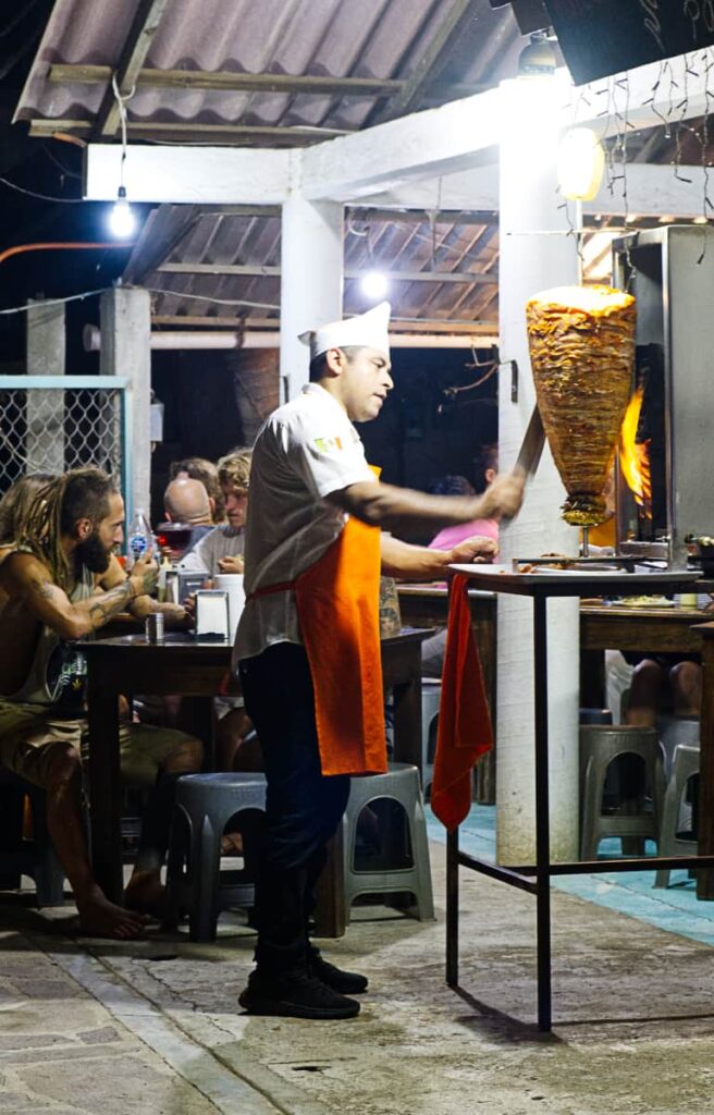 At the best restaurant for cheap tacos in Mazunte, a man wearing an apron slices pieces of al pastor from the spit of meat as it turns in front of a flame. Customers sit at tables in the background.