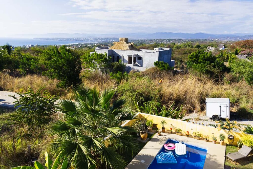 A pool is floats is shown at this coliving and hostel in Puerto Escondido. In the background is a concrete structure with palapa roof and sweeping views of the ocean and mountains in the background.