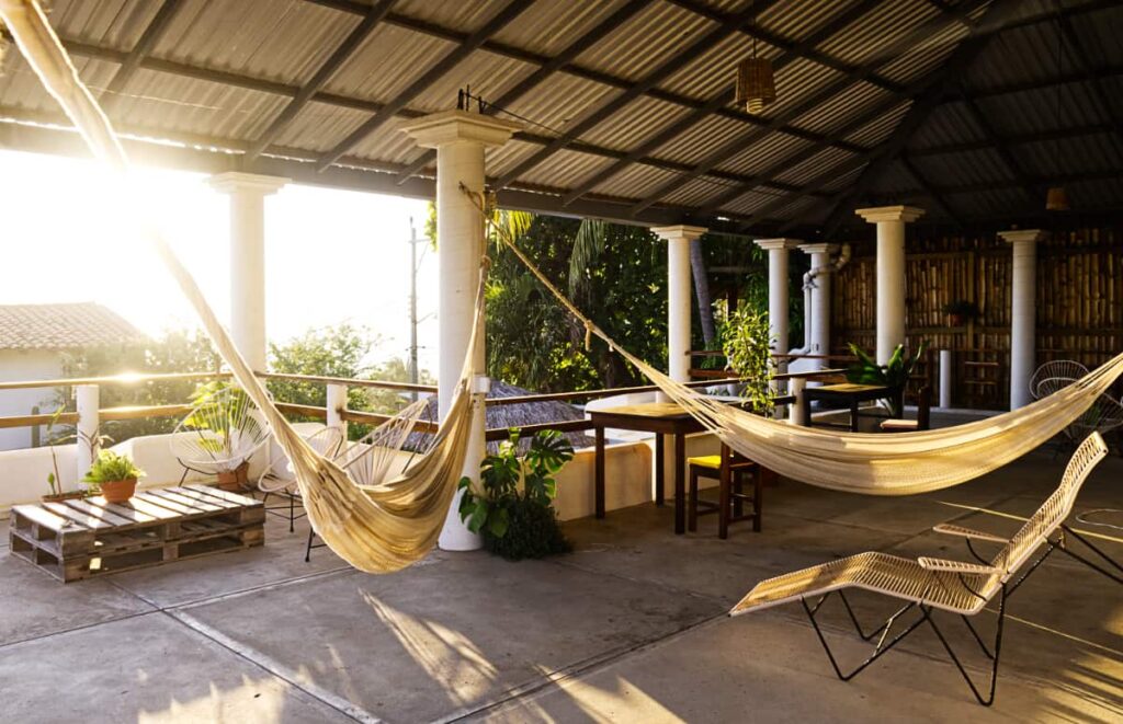 On the rooftop of Casa Flow Surf House and Coliving, two hammocks are suspended between white columns with the setting sun creating long shadows. There are other chairs and tables in the background.