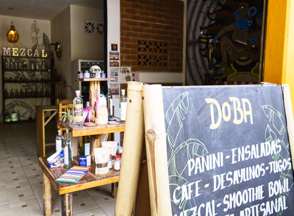 A three tiered table displays various goods for sale at Doba Restaurant Mazunte. In front is a sandwich board framed in bamboo that lists some of the specialities at Doba Restaurant. In the background is a metallic sign advertising mezcal with several shelves bottles below.