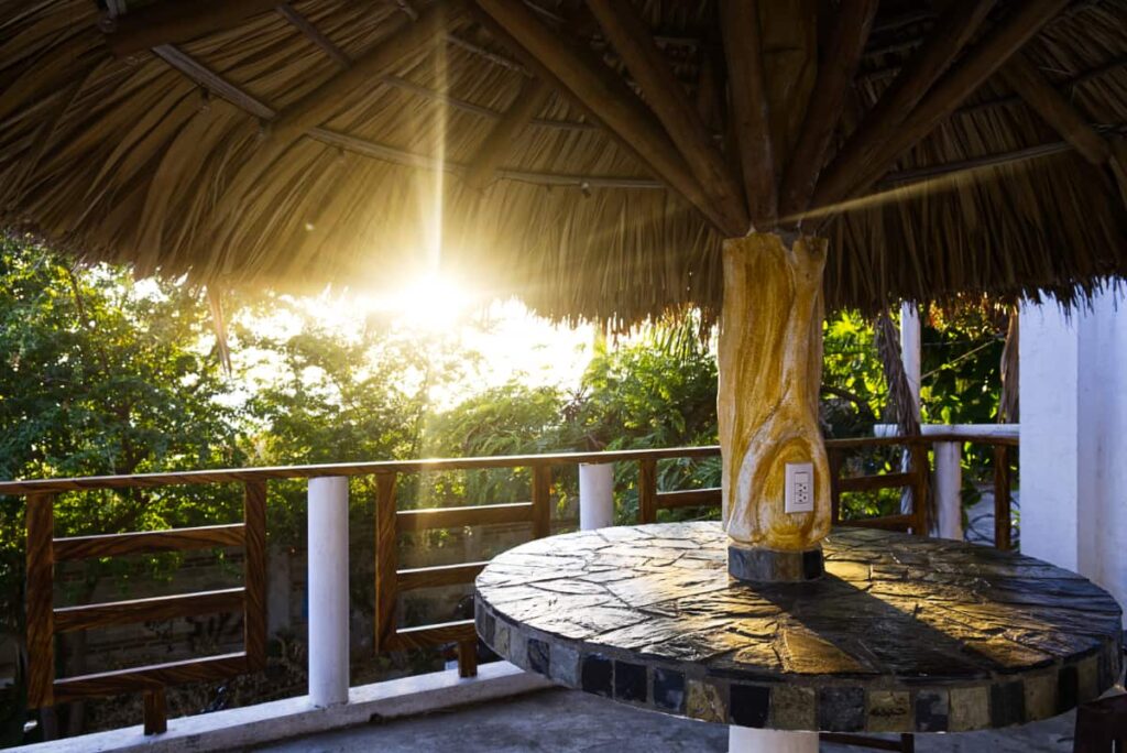 The setting sun cuts through the fronds of the palapa that covers a small table on the rooftop of Casa Flow Surf House and Coliving Puerto Escondido. The wooden table base includes a double outlet.