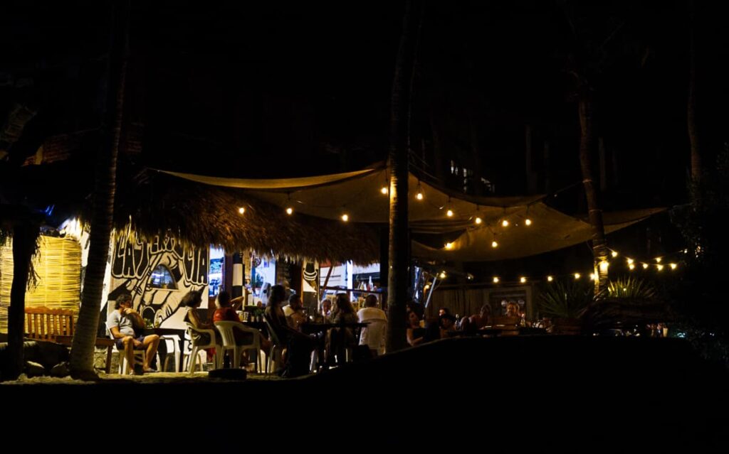 A night time view of one of the best beach restaurants in Mazunte, La Tertulia, shows tables full of people sitting under a fabric covering that is strung with lights.