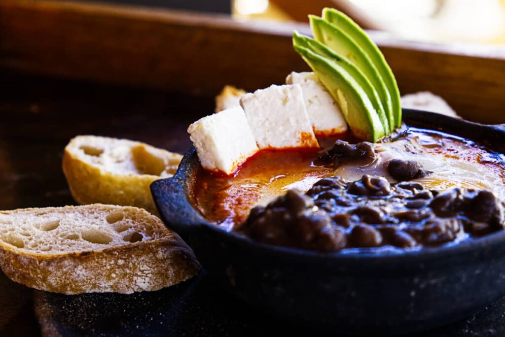 A close up view of one of the best breakfasts in Mazunte at Maralto Restaurant. The black metal bowl is full of a tomato sauce in eggs with black beans in the foreground, white panel cheese in the background with slices of avocado. On the side are several slices of homemade bread.