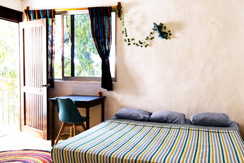 Inside a private room at El Mundito Coliving is a large bed made with striped sheets and three pillows. In front of the window next to the open wooden door is a small desk and a chair.