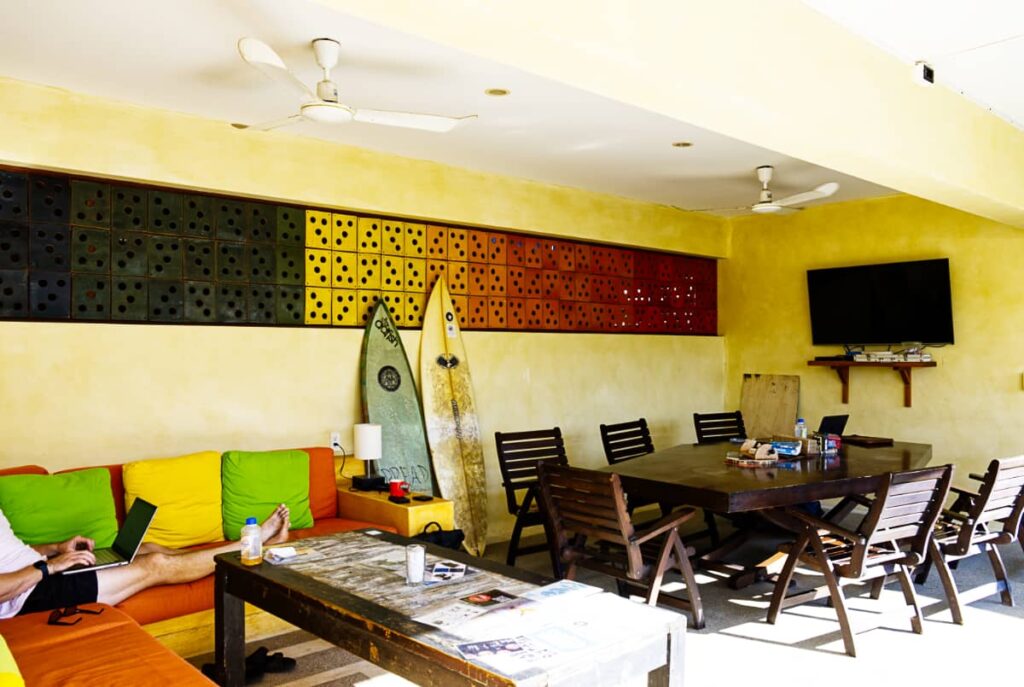 The coworking space at Bonobo coliving is a large wooden table with wooden chairs. A TV is hanging on one wall with two surfboards propped up on another. On the side is a yellow and orange couch. On the edge are a mans legs with a laptop resting on them.