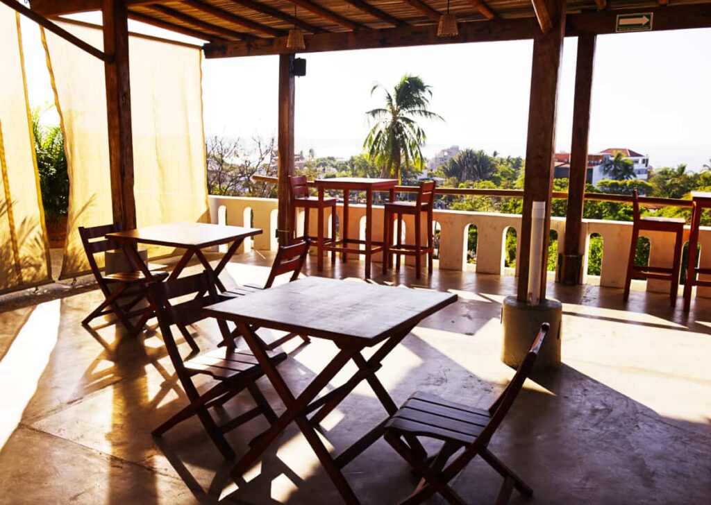 The shaded, open-air rooftop at Casa Losodeli features wooden tables and chairs with an ocean view. In the background is a tall palm tree.