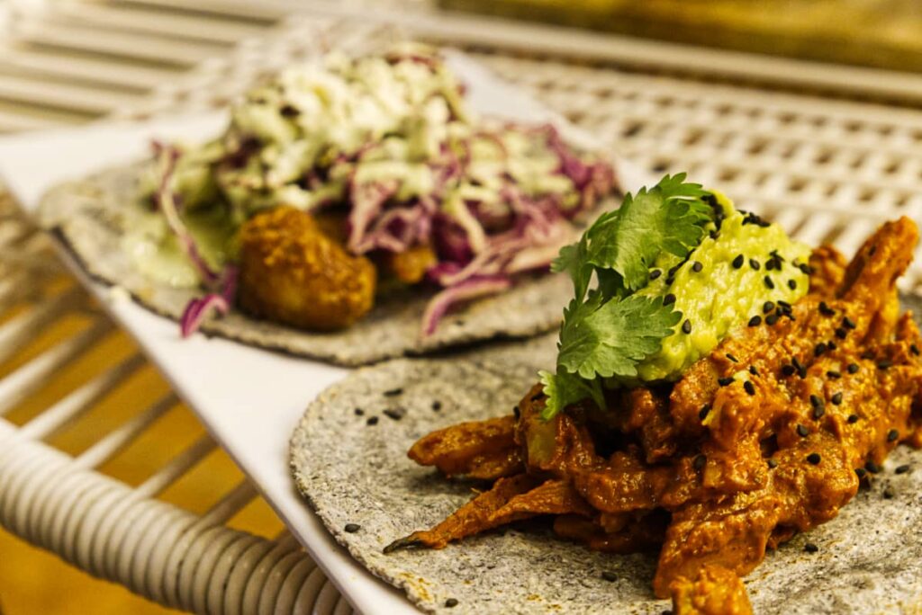 A plate of food at Tribu Restaurant in Mazunte features a vegetarian taco of sweet potato pastor topped with guacamole. In the blurred background is a fish taco topped with cabbage coleslaw.