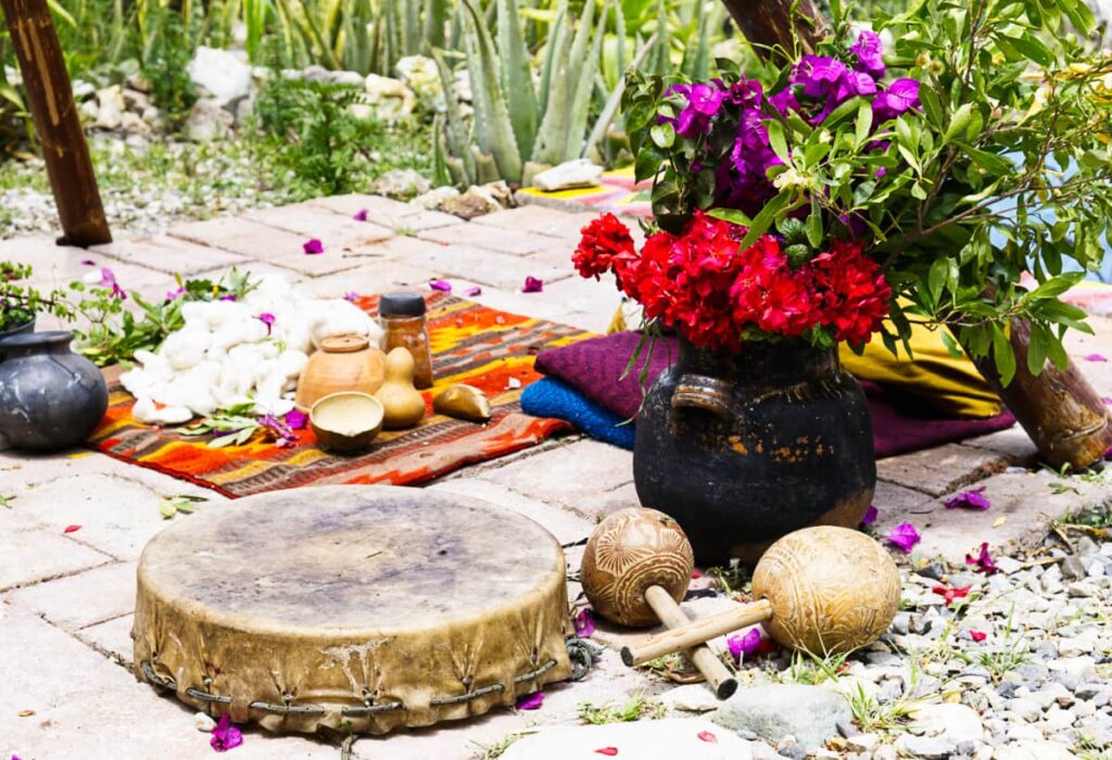 A round drum and other hand instruments sit on the ground with a pitcher of flowers. On a rug behind are several offerings for a Oaxaca temazcal ceremony.