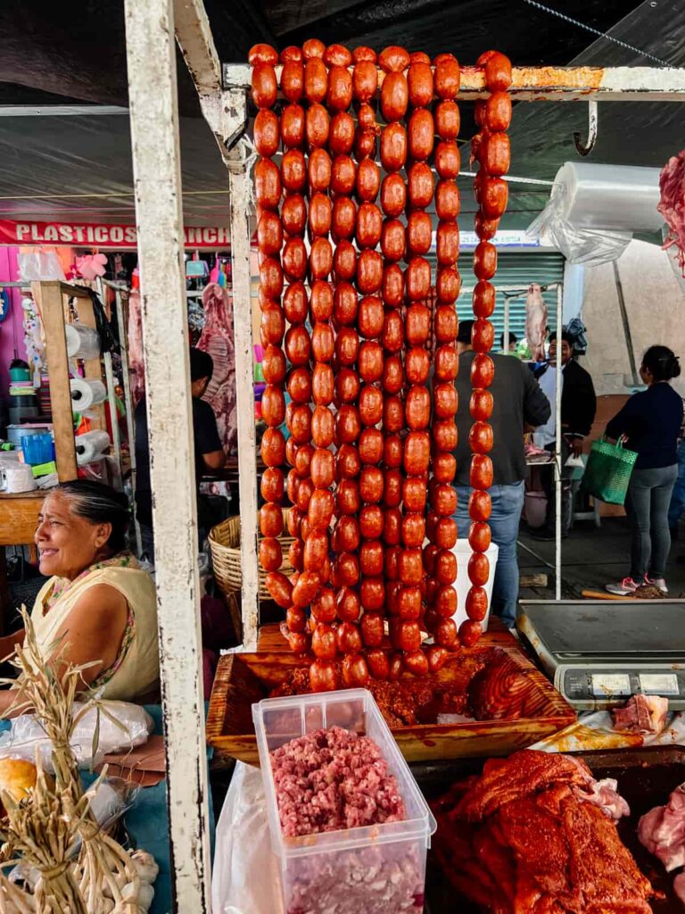 At the Abastos market in Oaxaca, long links of Oaxacan chorizo hang from the frame of the market stand. Other meat is piled in front with a jovial Oaxacan woman in the background.