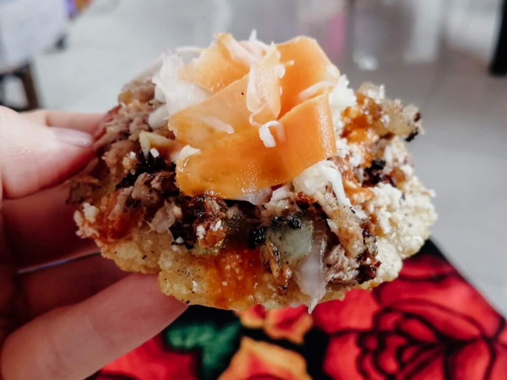 A hand holds a garnacha, a food from Oaxaca Ismto. Pictures is a small corn disk loaded with meat and salsa, topped with shredded cabbage and carrot.