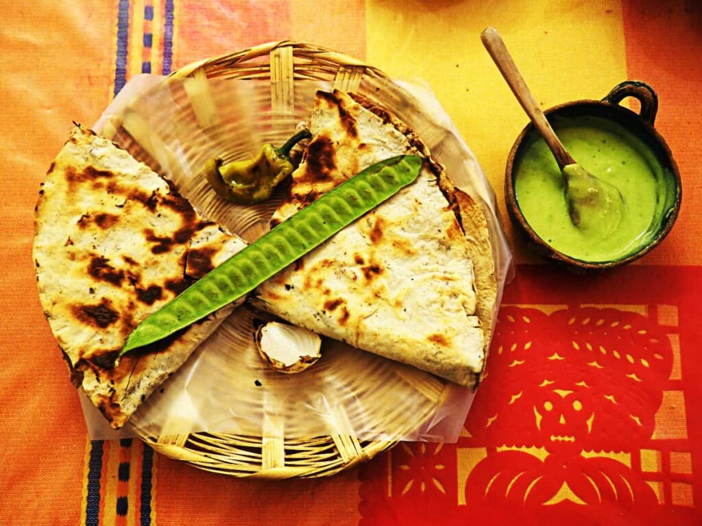 A overhead view of a tlayuda, the most popular Oaxacan food. It's served on a woven plate with the long green huaje pod placed on top at a diagonal. A small bowl of bright green salsa with a wooden spoon is on the side.