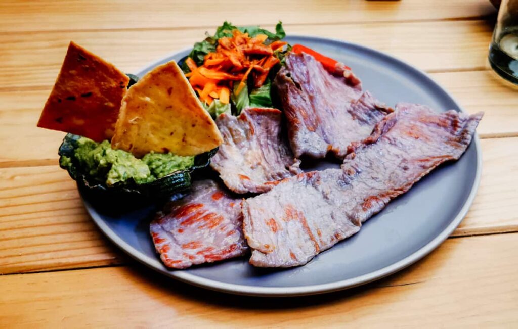 Pieces of grilled tasajo (thin cured beef) sit on a plate accompanied by guacamole and a small salad. Tasajo is a typical meat in Oaxaca, often served on a tlayuda.