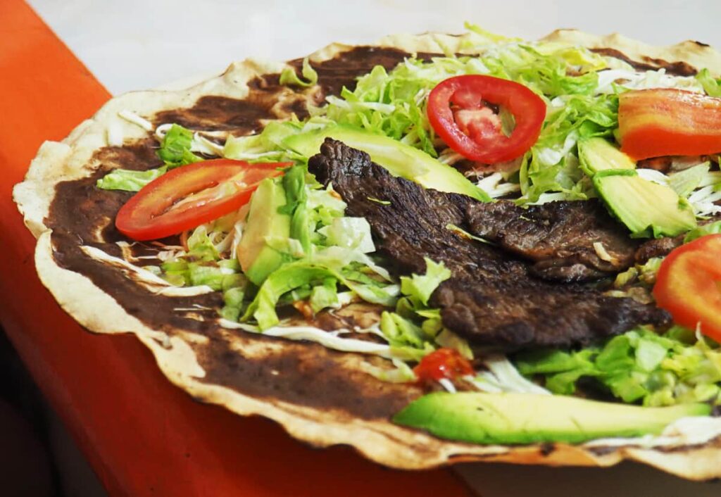 The most popular Oaxacan food, a tlayuda, shows a large tortilla spread with a bean paste, topped with cheese, shredded lettuce, avocado, tomato, and pieces of grilled tasajo.