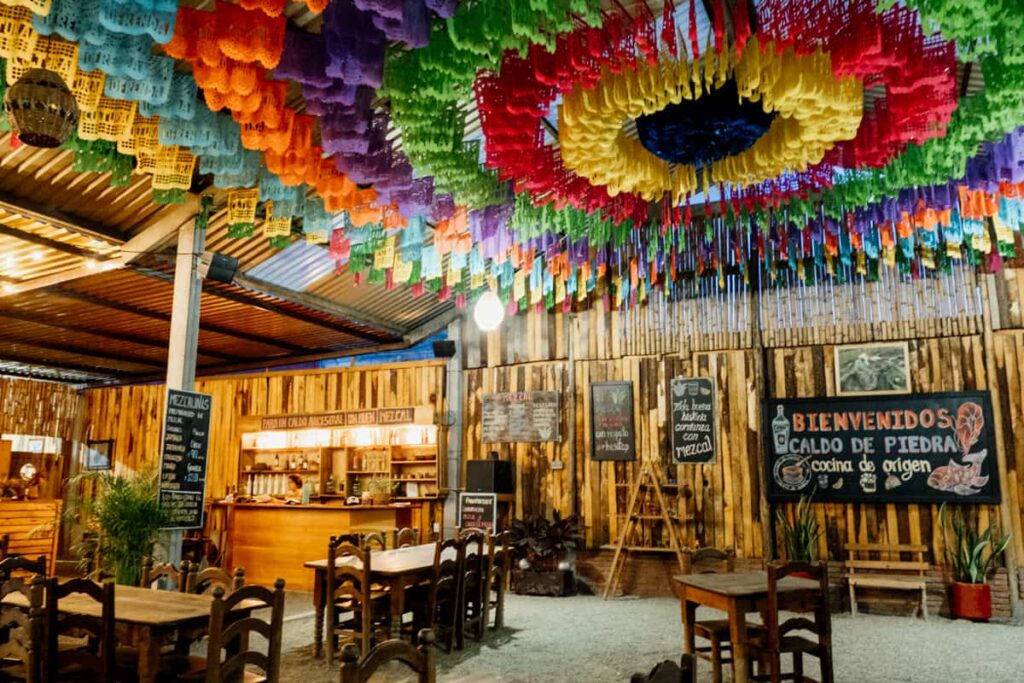 At Caldo de Piedra restaurant in Oaxaca, wooden tables and chairs sit under a tall roof decorated with a large papel picado, colorful paper in a circular pattern. On the walls are signs advertising drinks and mezcal as well as a welcome sign.