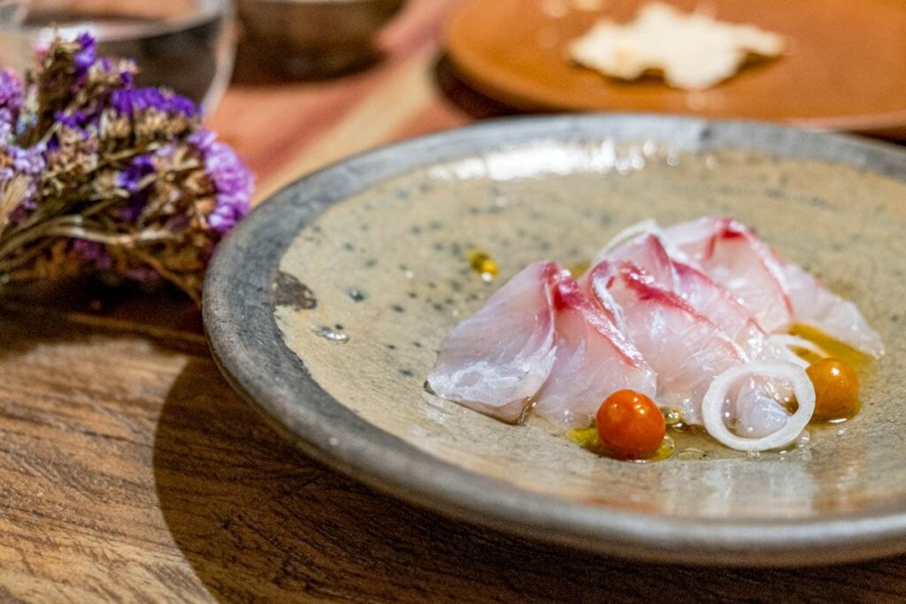 A close view of thinly sliced fish with cherry tomatoes in a light oil - one of the dishes from the tasting menu at Cataran restaurant in Oaxaca.