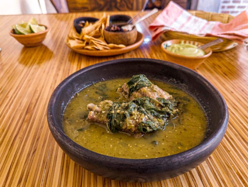 A round black bowl with ribs bathed in a green salsa sits on a table at Mook, one of the best restaurants in Oaxaca. In the background is a bowl of limes, a plate of chips with three bowls of salsa, a basket of tortillas wrapped in a towel, and a bowl of guacamole.