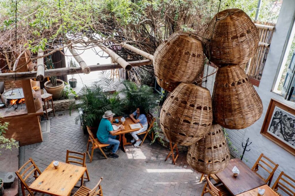 An overhead views of the downstairs seating at Luz de Lua in Huayapam Oaxaca shows a man an woman sitting at a wooden table surrounded by plants and some artwork hanging on the walls. In the background a comal is heated with a wood fire.