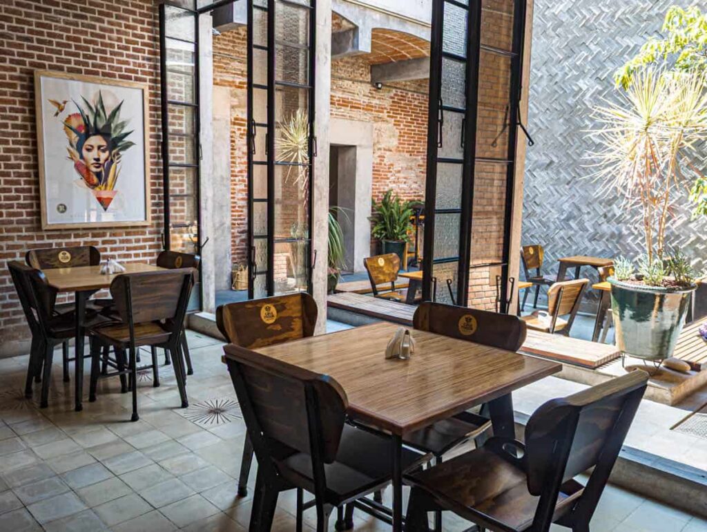 Wooden tables and chairs in the back dining room at Mook - one of the best restaurants in Oaxaca. Beyond the open doors is a small patio. On the wall hangs a piece of art that features a woman's face surrounded by corn and other ingredients.