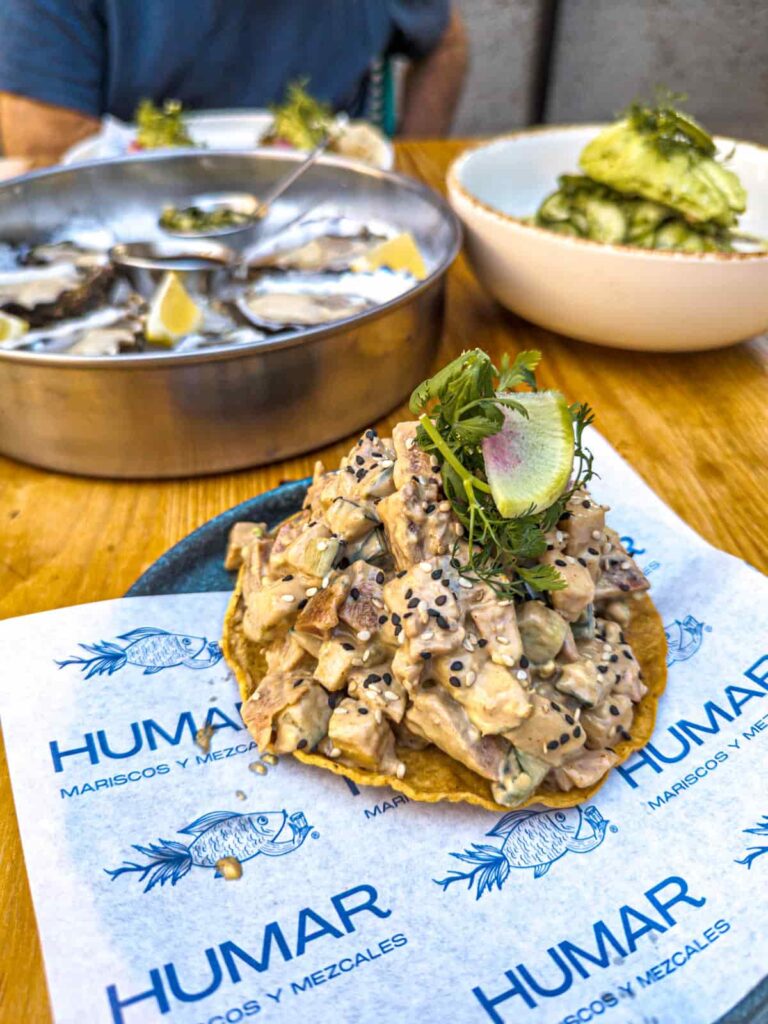 A tostada piled high with tuna and topped with greens on a plate lined with a paper that says Humar Mariscos y Mezcal. In the background are oysters on the half shell and aguachile in a large bowl.