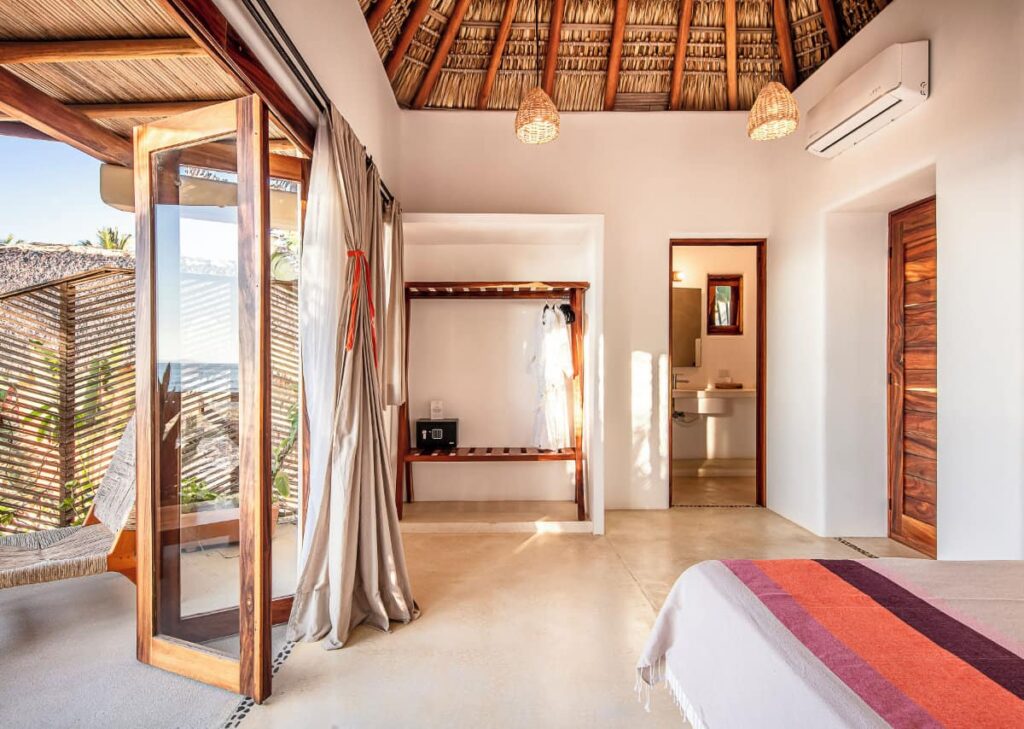 The all-white air-conditioned suite at Casa Kuaa with a thatched ceiling, connected bathroom, wooden accents and large glass doors that open to a private outdoor space.