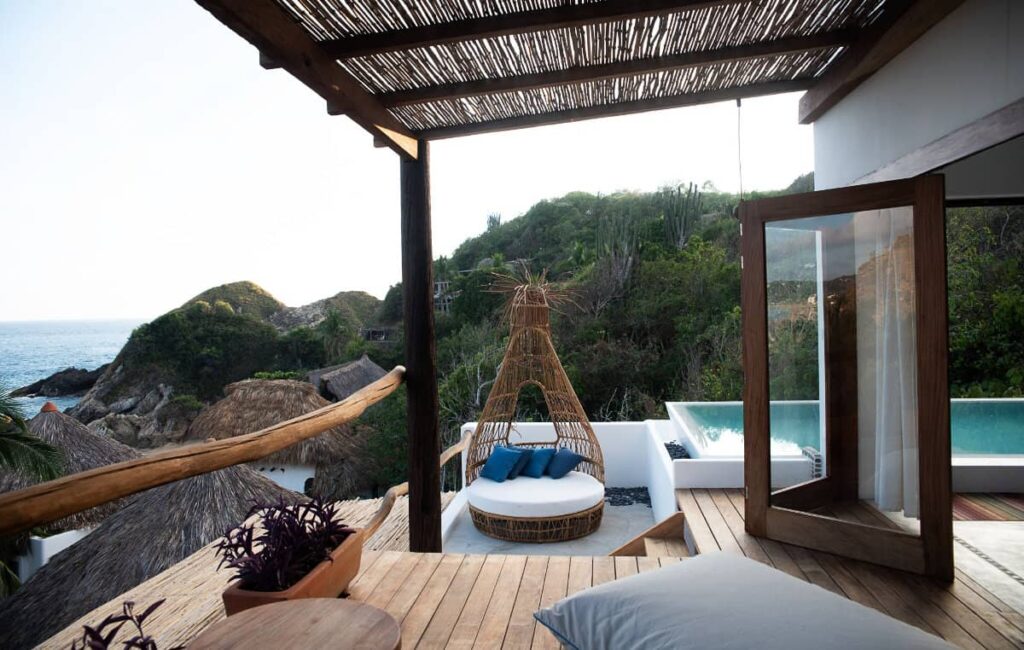 A balcony at El Alquimista, a beach resort in Oaxaca, with a lounger and an outdoor pool overlooking the lush greenery and partial ocean view.