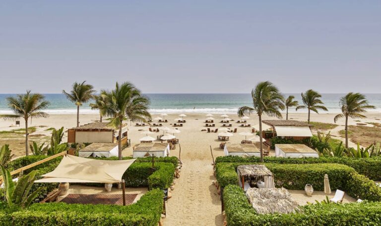 A view of one of the best beach resorts in Oaxaca features a sandy path leading to an ocean with lush green hedges and private cabanas with thatched roofs.