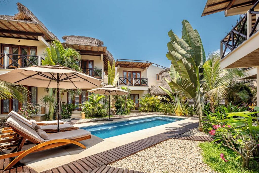 A view of a boutique hotel in Puerto Escondido with thatched roof, an outdoor pool area furnished with parasols, sunloungers and lush greenery.