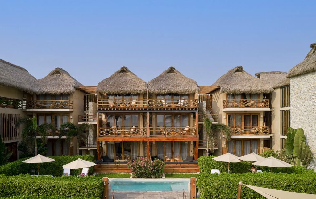 A view of Casa Joseph beachfront resort featuring thatched roof, balconies with private seating, an outdoor pool and lush green hedges.