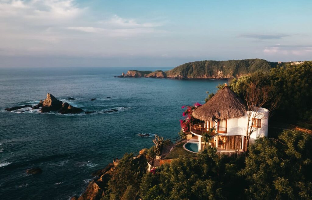 An overhead view of a white-washed ocean-side bungalow at ZOA with a thatched roof and a private swimming pool surrounded by lush greenery and outdoor space.