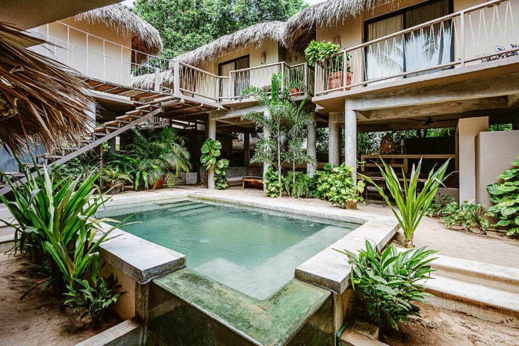 A view of the outdoor pool at Raiz Hotel Boutique surrounded by lush green spaces and rooms with thatched roofs.