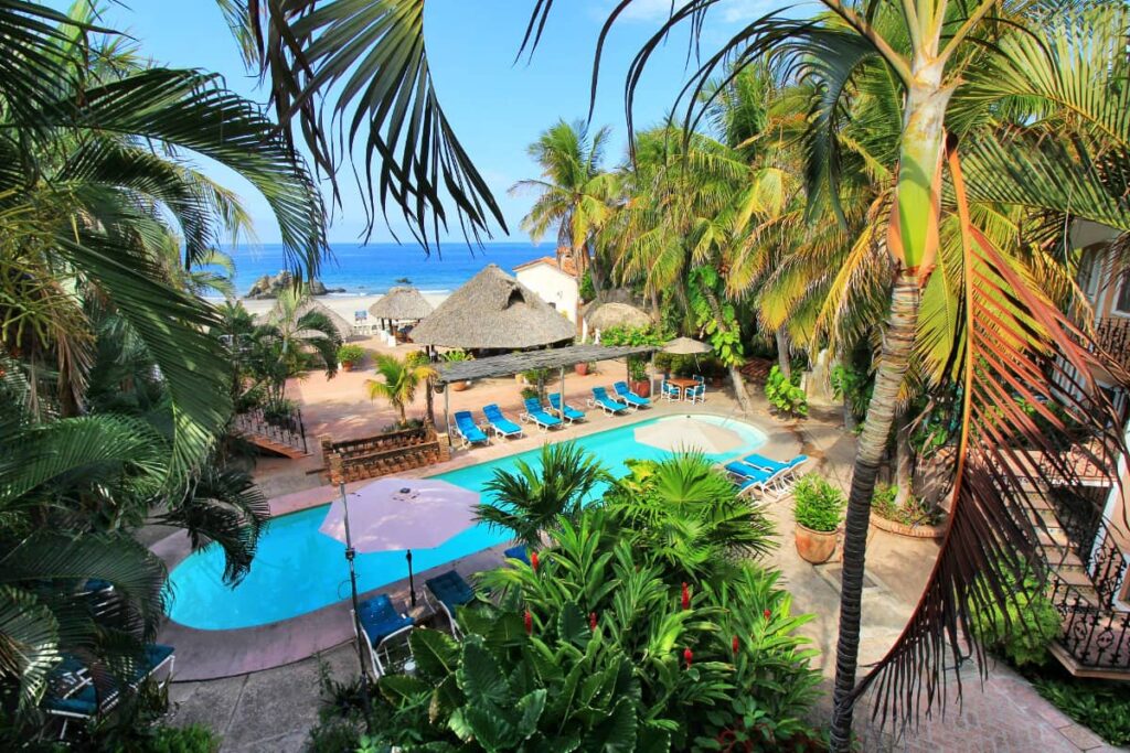 A pool view at Hotel Santa Fe featuring sunloungers and thatched roof spaces surrounded by palm trees, lush greenery and partial ocean view.